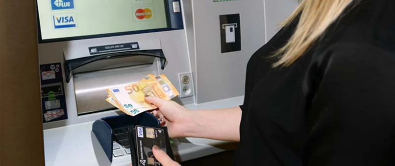 En kvinde står med euro sedler ude foran en pengeautomat.