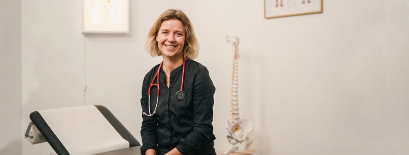 Kvindelig læge sidder med et stort smil på briksen i sin klinik