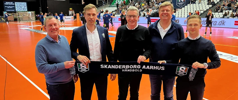 Fem mænd står på en håndboldbane og holder sammen et tørklæde med teksten "Skanderborg Aarhus Håndbold". De markere at Djurslands Bank indgår sponsorat med klubben.