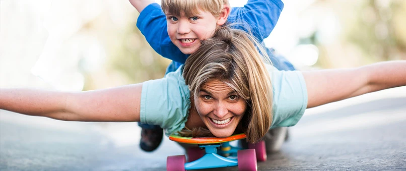 En kvinde ligger på et skatebord med armene ud til siden og smiler. Ovenpå hende ligger en lille dreng og smiler. 