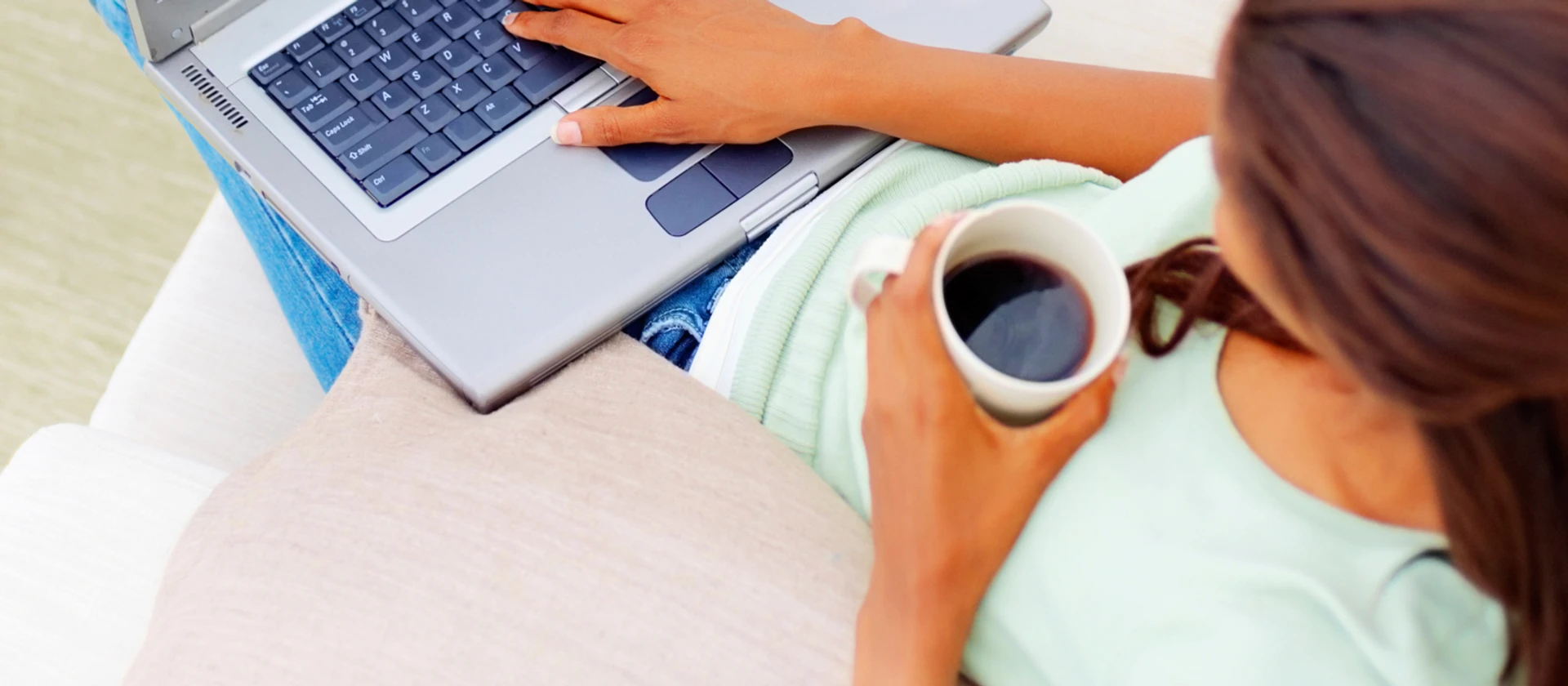 Kvinde sidder på sofa og drikker kaffe imens hun kigger på en bærbar computer.
