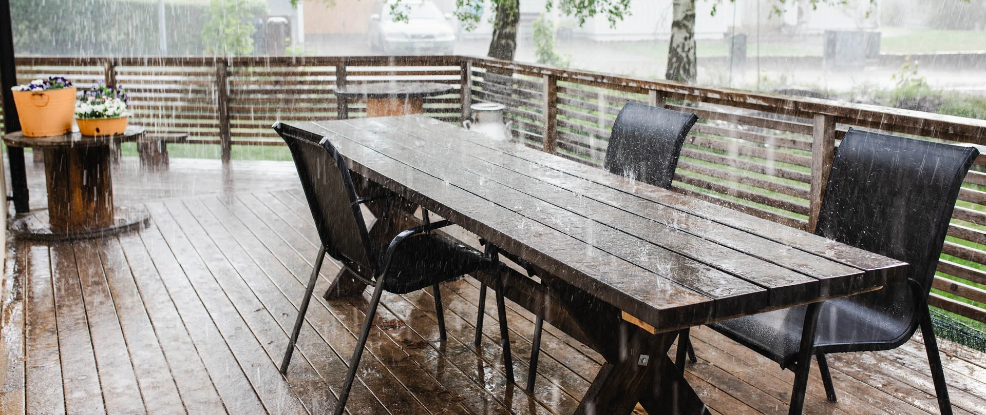 Voldsomt regnvejr på et terrassebord og nogle terrassestole der står på en træterrasse.