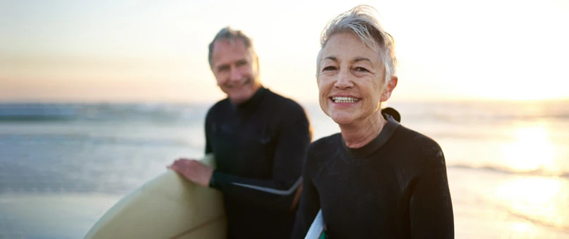 Ældre par på strand i solnedgang iført våddragter med et surfboard
