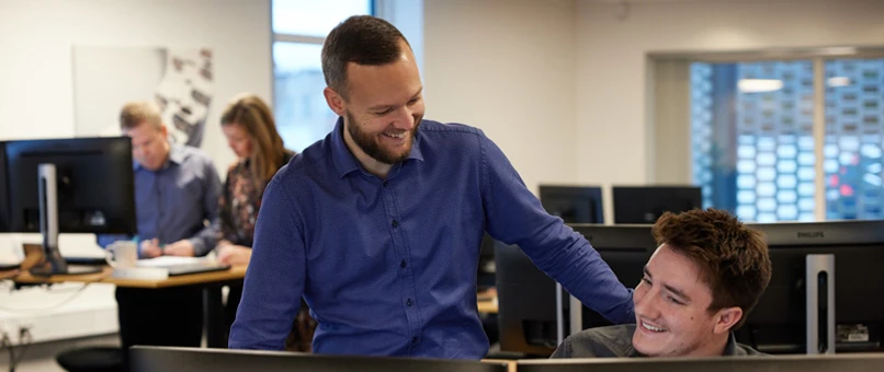 To Djurslands Bank medarbejdere der smiler og griner. Den ene sidder på en stol bag skrivebordet og kigger på en computer, den anden står ved siden af og kigger på sin kollega, imens han griner og har hånden på skulderen af kollegaen.